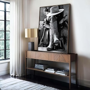 May include: Black and white framed print of a cowboy seated on a saddle, wearing a hat and detailed cowboy boots. The artwork is displayed on a wooden console table with a lamp and books, set against a window with white curtains.