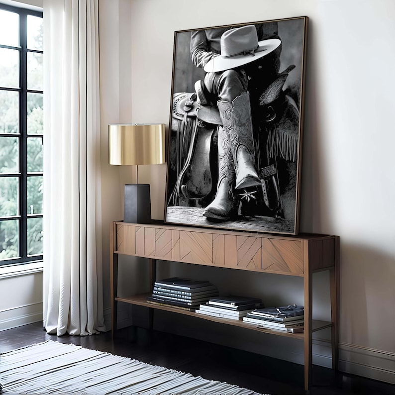 May include: Black and white framed print of a cowboy seated on a saddle, wearing a hat and detailed cowboy boots. The artwork is displayed on a wooden console table with a lamp and books, set against a window with white curtains.