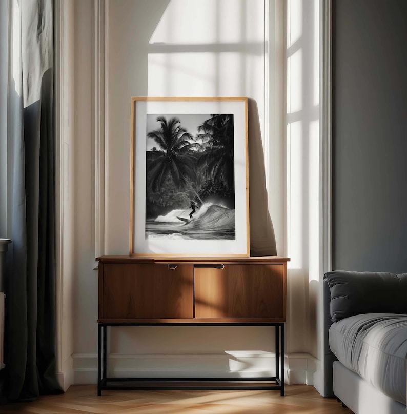 May include: A framed black and white photograph of a surfer riding a wave, set against a backdrop of palm trees. The photograph is displayed on a wooden cabinet with a black metal frame, in a room with natural light.