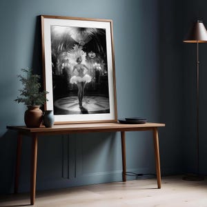 May include: Black and white framed print of a showgirl in a feathered headdress and tutu, standing on a circular stage. The print is displayed on a wooden table with a small plant and vase.