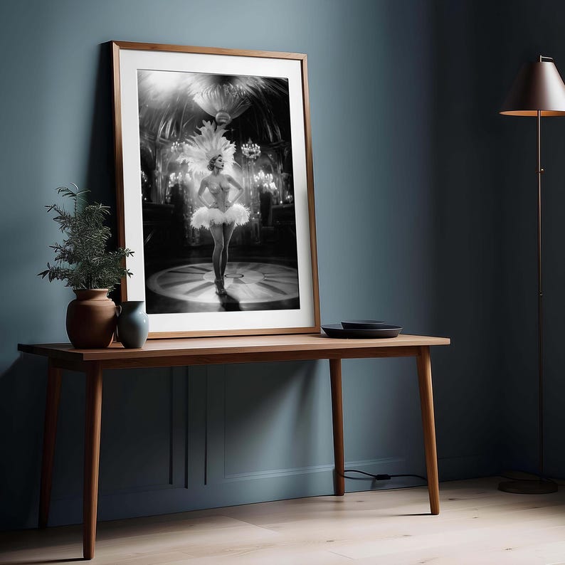 May include: Black and white framed print of a showgirl in a feathered headdress and tutu, standing on a circular stage. The print is displayed on a wooden table with a small plant and vase.