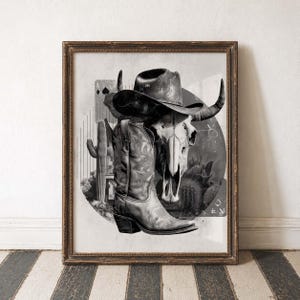 Op de afbeelding: Ingelijste zwart-wit print met een cowboyhoed, stierschedel en een cowboylaars. Het kunstwerk heeft een woestijnthema met cactussen en een vintage esthetiek. De lijst is donkerbruin.