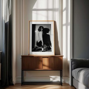 K&ouml;nnte beinhalten: Gerahmte Schwarz-Wei&szlig;-Fotografie einer knienden Person, die eine Weinflasche h&auml;lt, mit einem Weinglas. Die Fotografie wird auf einem Holzschrank mit schwarzem Metallrahmen in einem Raum mit nat&uuml;rlichem Licht ausgestellt.