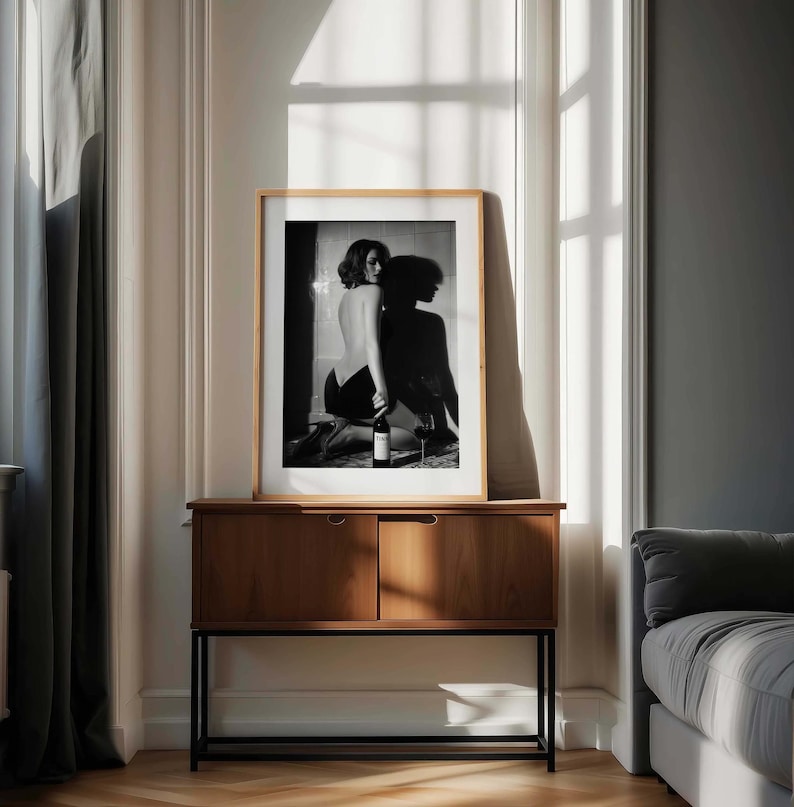 K&ouml;nnte beinhalten: Gerahmte Schwarz-Wei&szlig;-Fotografie einer knienden Person, die eine Weinflasche h&auml;lt, mit einem Weinglas. Die Fotografie wird auf einem Holzschrank mit schwarzem Metallrahmen in einem Raum mit nat&uuml;rlichem Licht ausgestellt.