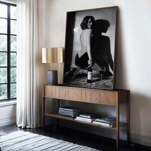 K&ouml;nnte beinhalten: Schwarz-wei&szlig; gerahmtes Foto einer Frau mit Weinflasche und Glas, auf einem Holztisch. Eine goldene Lampe und B&uuml;cher stehen auf dem Tisch. Im Hintergrund ein Fenster mit wei&szlig;en Vorh&auml;ngen.
