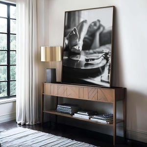 K&ouml;nnte beinhalten: Ein schwarz-wei&szlig; gerahmtes Foto einer Person in der N&auml;he eines Plattenspielers. Eine goldene Lampe mit schwarzem Sockel steht auf einer Holzkonsolentisch. B&uuml;cher und Schallplatten befinden sich auf den Regalen des Tisches. Im Hintergrund befindet sich ein Fenster mit wei&szlig;en Vorh&auml;ngen.