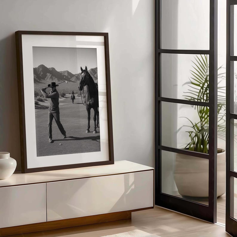 May include: Black and white framed print featuring a golfer swinging a club on a desert golf course. A horse stands nearby. The print is displayed on a white cabinet with a wooden base, next to a glass door and a potted plant.