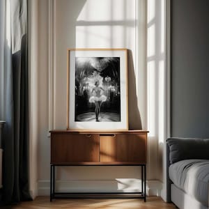 May include: Black and white framed print of a showgirl in a feathered headdress and tutu, standing on a stage. The print is displayed on a dark wood cabinet with a black metal base, set against a neutral wall and window.