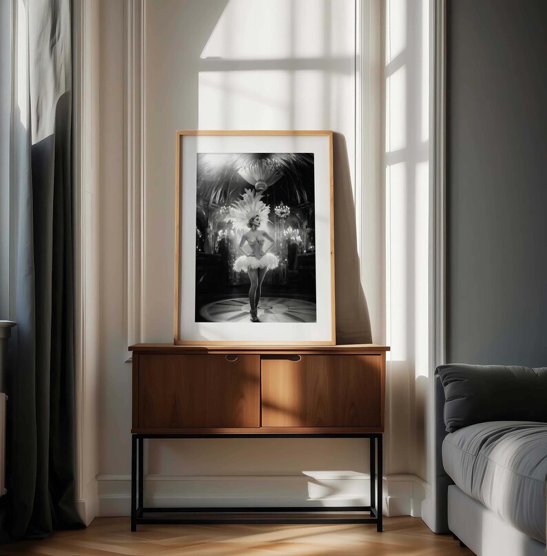 May include: Black and white framed print of a showgirl in a feathered headdress and tutu, standing on a stage. The print is displayed on a dark wood cabinet with a black metal base, set against a neutral wall and window.