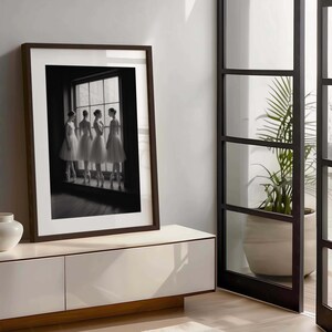 May include: A framed black and white photograph of four ballerinas in white tutus and pointe shoes, standing by a window. The photograph is in a dark brown frame, displayed on a white cabinet.