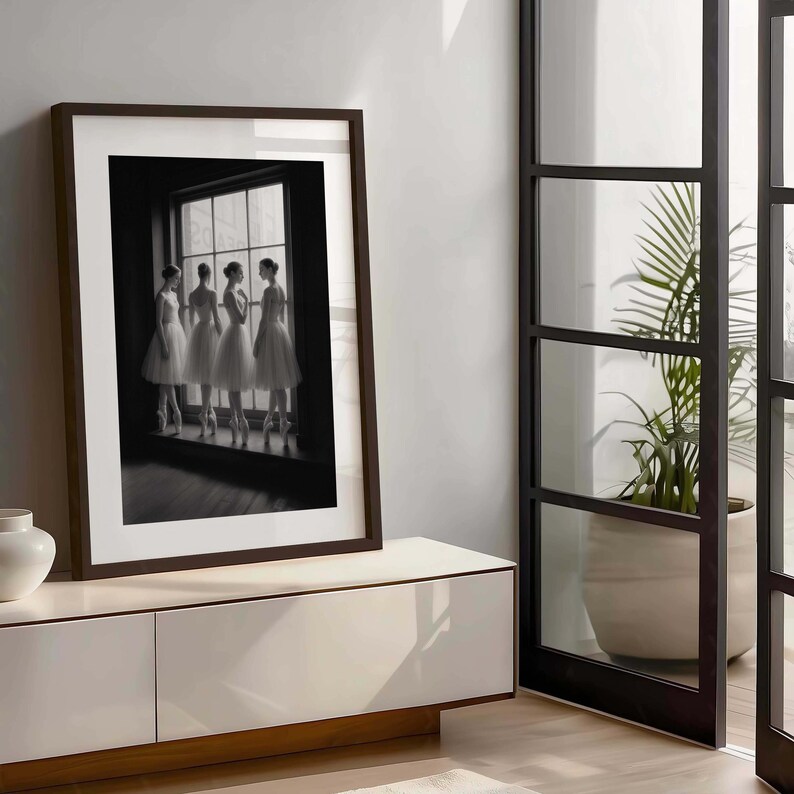 May include: A framed black and white photograph of four ballerinas in white tutus and pointe shoes, standing by a window. The photograph is in a dark brown frame, displayed on a white cabinet.