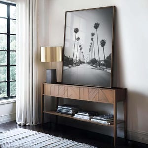 May include: A black and white framed photograph of a street lined with palm trees, displayed on a wooden console table. A gold-toned lamp with a black base sits on the table. Books and decorative items are on the lower shelf.