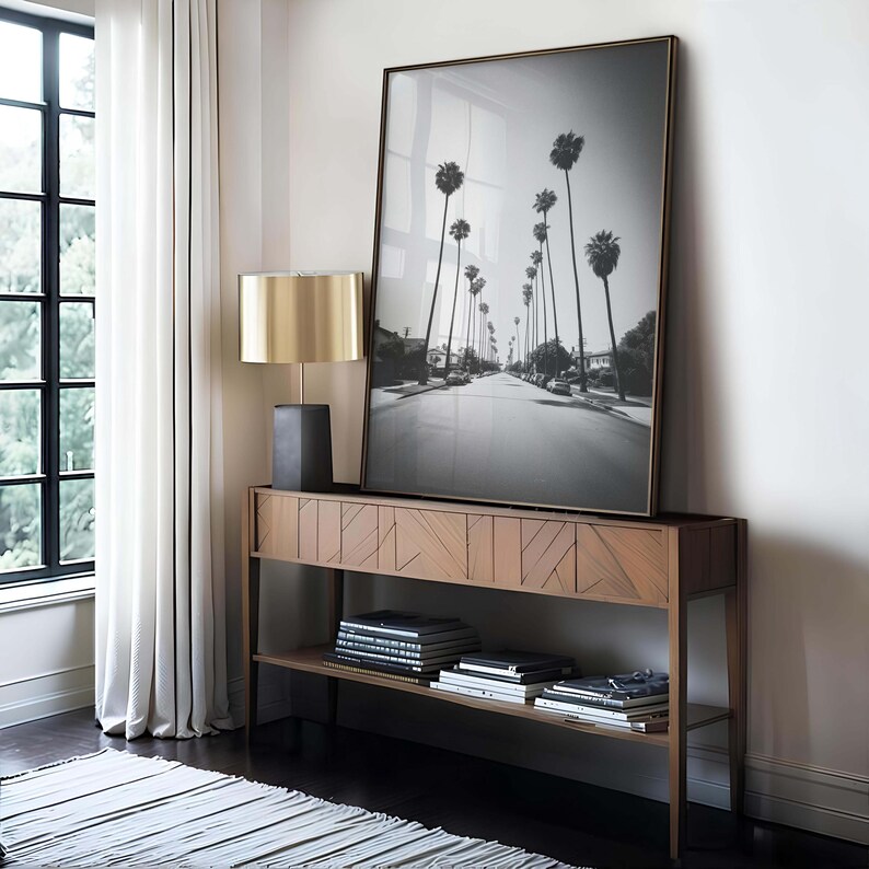 May include: A black and white framed photograph of a street lined with palm trees, displayed on a wooden console table. A gold-toned lamp with a black base sits on the table. Books and decorative items are on the lower shelf.