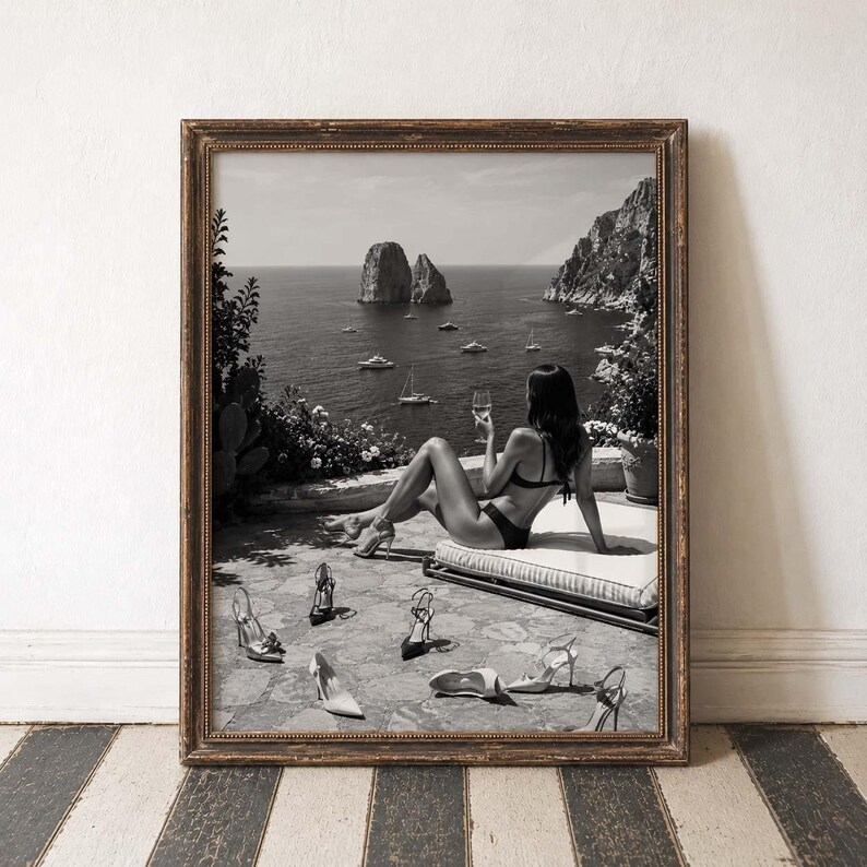 May include: A framed black and white wall art print depicting a person in a bikini sitting on a lounge chair on a terrace overlooking the sea with rock formations and boats. Several pairs of high-heeled shoes are scattered on the stone floor.