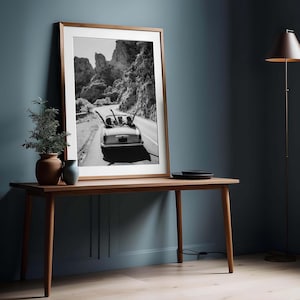 Puede incluir: Impresi&oacute;n en blanco y negro enmarcada de un coche descapotable cl&aacute;sico en una carretera de monta&ntilde;a. La impresi&oacute;n se muestra sobre una mesa de madera, con una planta y un jarr&oacute;n.