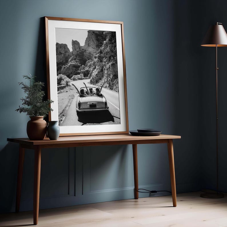 Puede incluir: Impresi&oacute;n en blanco y negro enmarcada de un coche descapotable cl&aacute;sico en una carretera de monta&ntilde;a. La impresi&oacute;n se muestra sobre una mesa de madera, con una planta y un jarr&oacute;n.