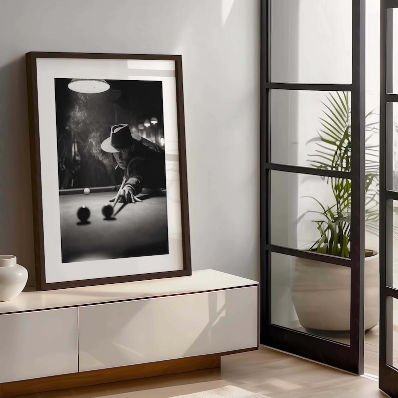 May include: Monochrome framed print depicting a person playing pool, wearing a hat and smoking. Two pool balls are in the foreground. The print is displayed on a white cabinet with a wooden base.