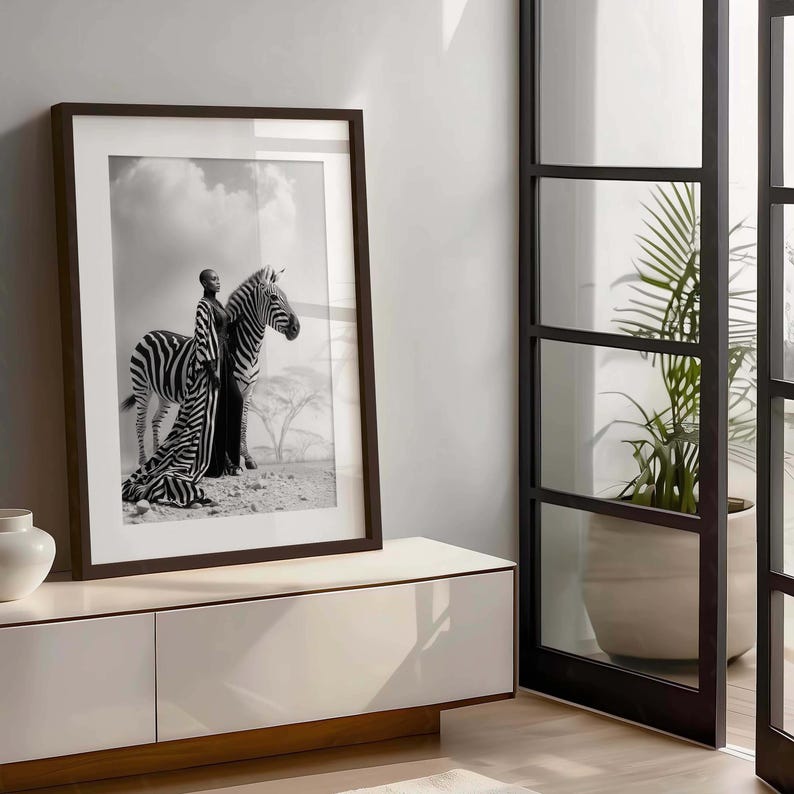 May include: Monochrome framed print depicting a person in a striped outfit alongside a zebra. The image is set in a desert landscape with a cloudy sky. The dark brown frame complements the black and white photograph.