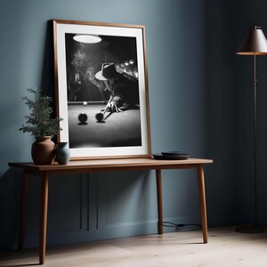 May include: A framed monochrome photograph of a person playing snooker, smoking a cigarette. The photograph is in a wooden frame and rests on a wooden table. A small potted plant and a vase are on the table, with a floor lamp to the right.