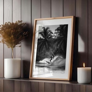 May include: Black and white framed print of a surfer riding a wave. The image features a surfer silhouetted against a wave with palm trees in the background. The frame is a light wood color, and the print is displayed on a shelf with a candle and a plant.
