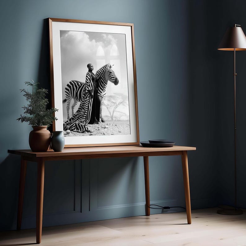 May include: Monochrome framed print depicting a person and a zebra in a desert setting. The artwork is displayed on a wooden table against a dark blue wall. A floor lamp and decorative items complete the scene.