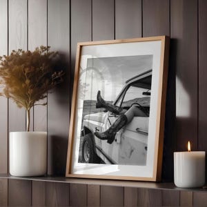 Pu&ograve; includere: Fotografia incorniciata in bianco e nero di una persona con stivali da cowboy che si sporge da un camion bianco. La fotografia &egrave; esposta su una mensola di legno accanto a una candela bianca e una pianta in vaso.