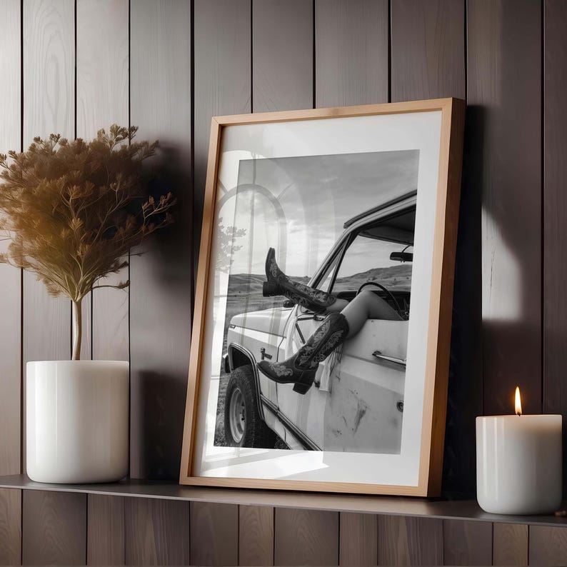 Pu&ograve; includere: Fotografia incorniciata in bianco e nero di una persona con stivali da cowboy che si sporge da un camion bianco. La fotografia &egrave; esposta su una mensola di legno accanto a una candela bianca e una pianta in vaso.