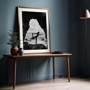 K&ouml;nnte beinhalten: Gerahmtes Schwarz-Wei&szlig;-Foto einer Person, die eine Yoga-Pose an einem felsigen Ufer ausf&uuml;hrt, eingerahmt von einem nat&uuml;rlichen Torbogen. Der Rahmen ist warmbraun und das Bild wird auf einem Holztisch pr&auml;sentiert. Eine kleine Pflanze in einem Terrakottatopf und eine blaue Vase stehen auf dem Tisch.