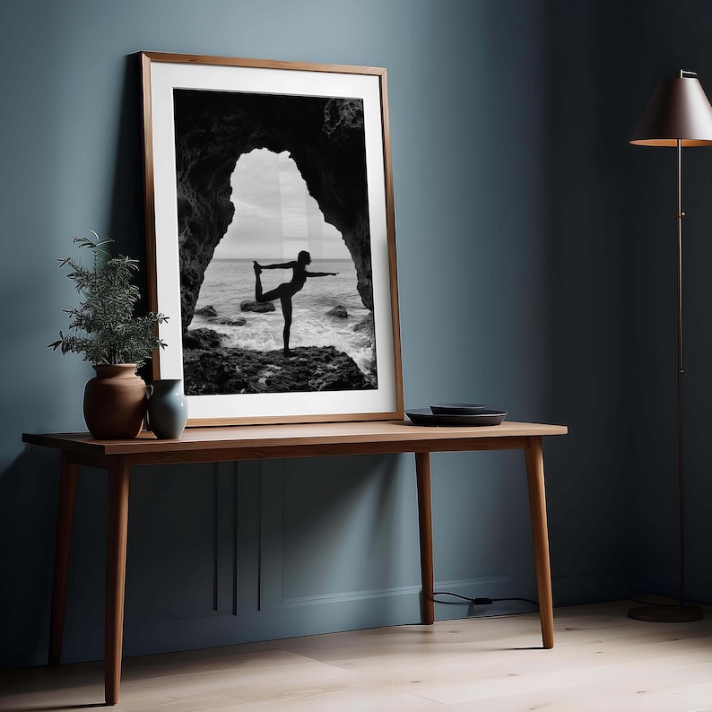 K&ouml;nnte beinhalten: Gerahmtes Schwarz-Wei&szlig;-Foto einer Person, die eine Yoga-Pose an einem felsigen Ufer ausf&uuml;hrt, eingerahmt von einem nat&uuml;rlichen Torbogen. Der Rahmen ist warmbraun und das Bild wird auf einem Holztisch pr&auml;sentiert. Eine kleine Pflanze in einem Terrakottatopf und eine blaue Vase stehen auf dem Tisch.
