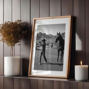 May include: Black and white framed print of a golfer swinging a club on a golf course, with a horse and rider in the background. The print is in a light wood frame, displayed on a shelf with a candle and a decorative plant.