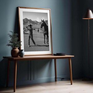 May include: A framed black and white photograph of a golfer swinging a club on a golf course, with a horse and rider in the background. The photograph is displayed on a wooden table against a blue wall.