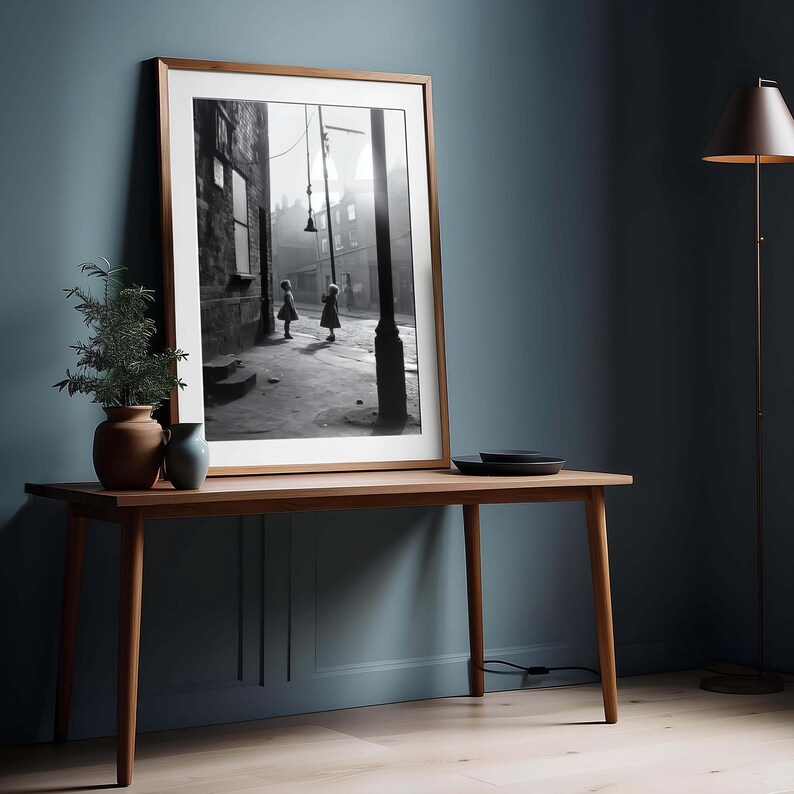 May include: Black and white photograph of two children walking down a narrow street. The photo is framed in a wooden frame and is displayed on a wooden table.
