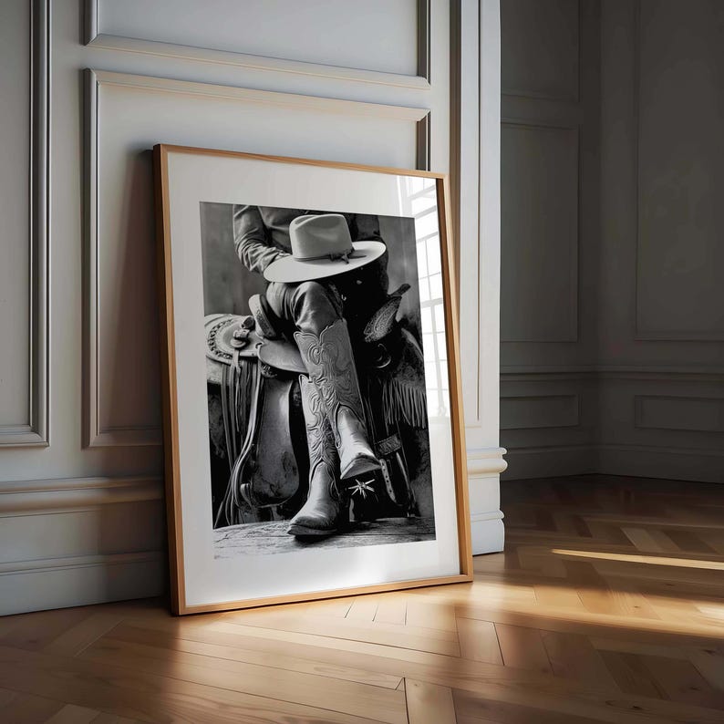 May include: Black and white framed print of a cowboy scene. The image features a person seated on a saddle, wearing cowboy boots and a hat. The boots have decorative stitching. The print is leaning against a wall with wooden flooring.