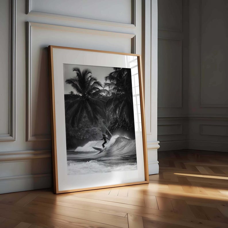 May include: Black and white framed print of a surfer riding a wave. The image features a surfer silhouetted against the wave, with palm trees in the background. The frame is a light wood color, and the print is leaning against a wall.