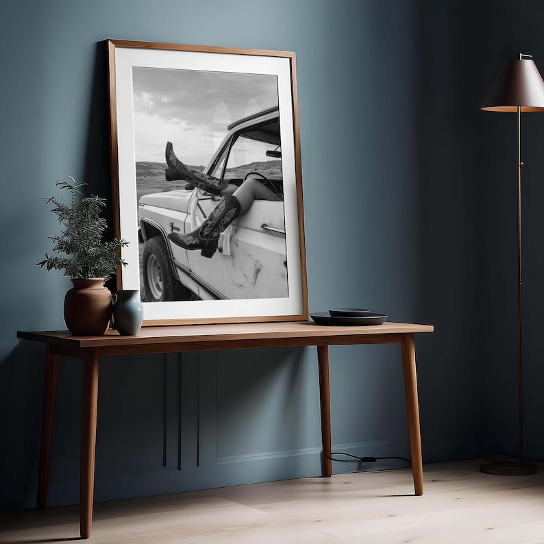 Pu&ograve; includere: Fotografia in bianco e nero incorniciata di stivali da cowboy e gambe che sporgono da un veicolo bianco. La fotografia &egrave; esposta su un tavolo di legno contro un muro blu. Gli stivali hanno un motivo decorativo.