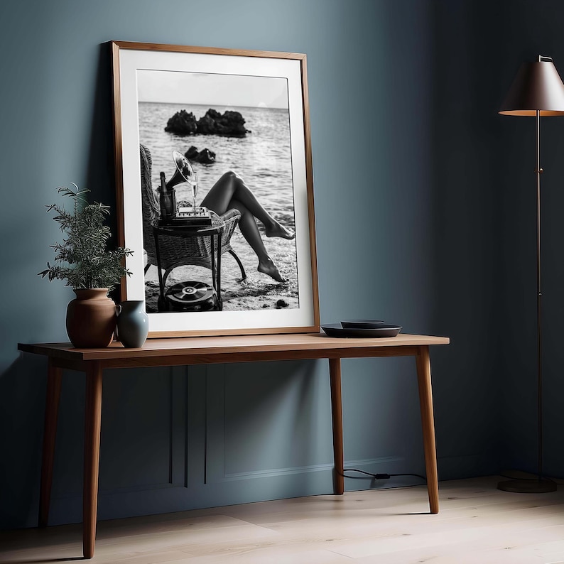 Pode incluir: Impress&atilde;o emoldurada em preto e branco de uma mulher relaxando em uma praia com um toca-discos vintage. A impress&atilde;o &eacute; exibida em uma mesa de madeira com uma pequena planta e um vaso.