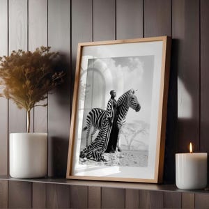 May include: A framed monochrome art print showing a person in a long dress standing beside a zebra. The print is in a light wood frame, displayed on a shelf with a candle and a potted plant.