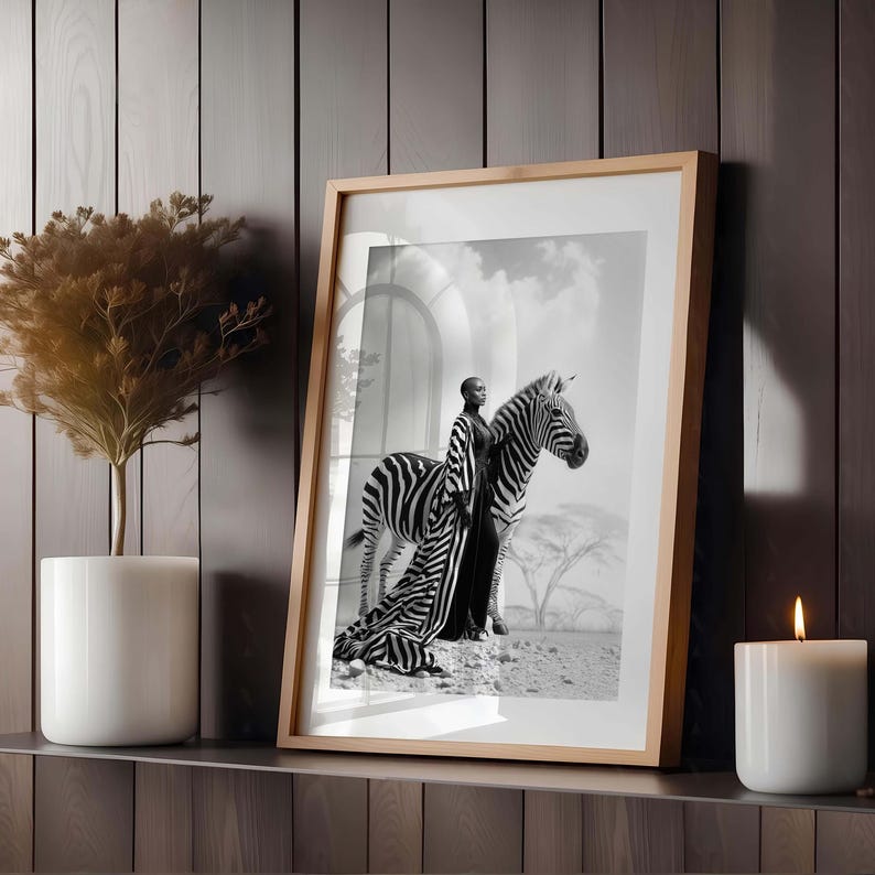 May include: A framed monochrome art print showing a person in a long dress standing beside a zebra. The print is in a light wood frame, displayed on a shelf with a candle and a potted plant.