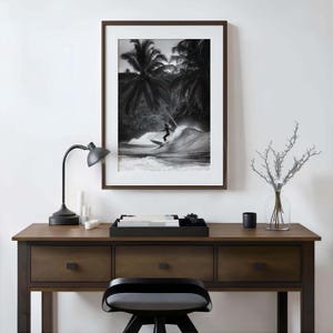 May include: Black and white framed print of a surfer riding a wave. The image is set against a backdrop of palm trees. The print is displayed on a dark wood desk with a black lamp and a decorative tray.