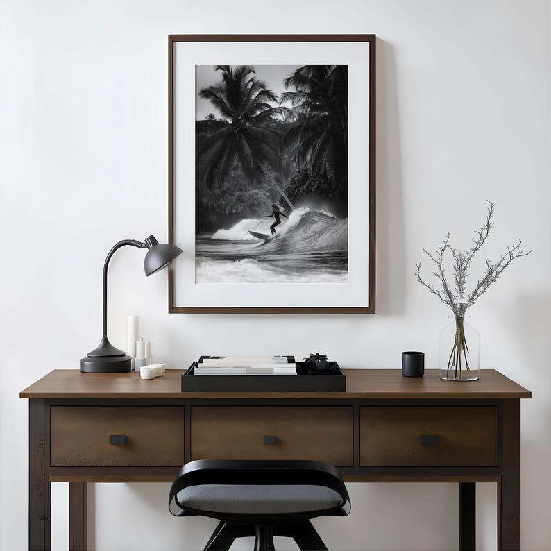 May include: Black and white framed print of a surfer riding a wave. The image is set against a backdrop of palm trees. The print is displayed on a dark wood desk with a black lamp and a decorative tray.