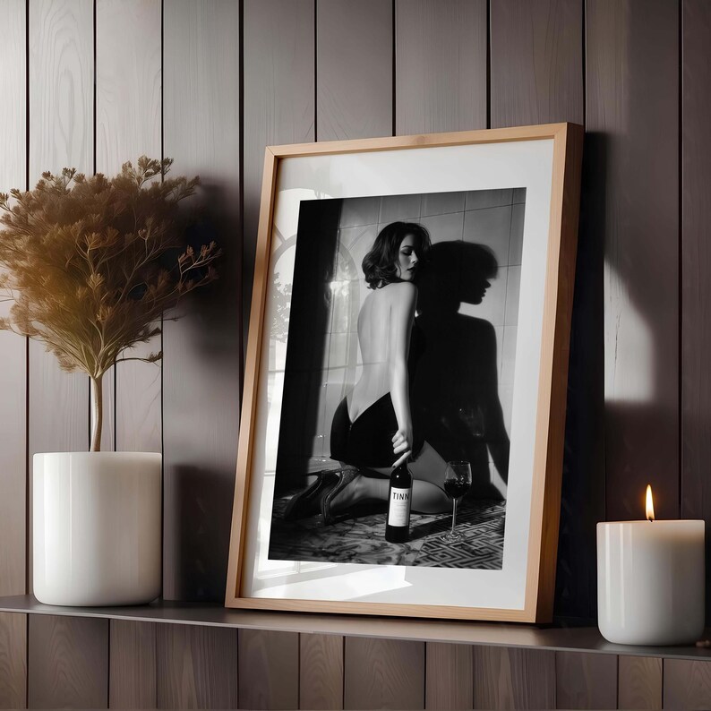 K&ouml;nnte beinhalten: Gerahmte Schwarz-Wei&szlig;-Fotografie einer knienden Frau mit einer Weinflasche und einem Weinglas. Das Foto befindet sich in einem hellen Holzrahmen und wird auf einem Regal mit einer wei&szlig;en Kerze und einer Topfpflanze ausgestellt.