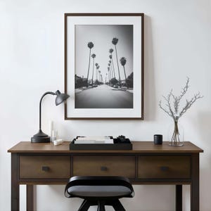 May include: A black and white framed print of a street lined with palm trees hangs above a wooden desk. A black desk lamp, candles, a tray, and a vase with branches are on the desk. A black chair is in front of the desk.