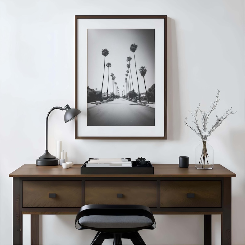May include: A black and white framed print of a street lined with palm trees hangs above a wooden desk. A black desk lamp, candles, a tray, and a vase with branches are on the desk. A black chair is in front of the desk.