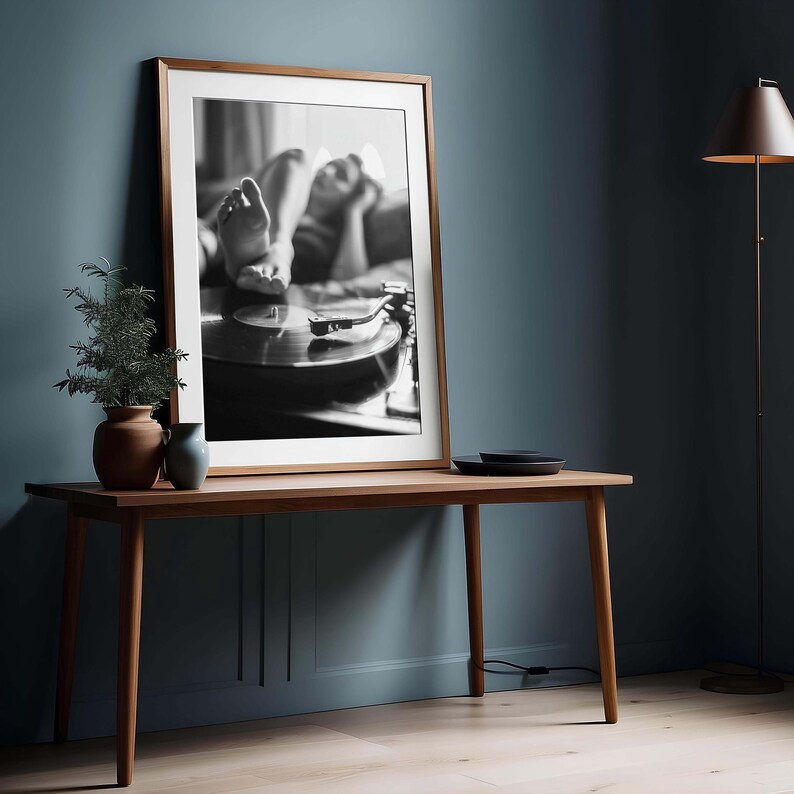 K&ouml;nnte beinhalten: Gerahmtes Schwarz-Wei&szlig;-Foto einer Person, die sich in der N&auml;he eines Plattenspielers entspannt. Das Foto wird auf einem Holztisch pr&auml;sentiert, mit einer kleinen Pflanze und einer Vase links. Rechts steht eine Stehlampe.