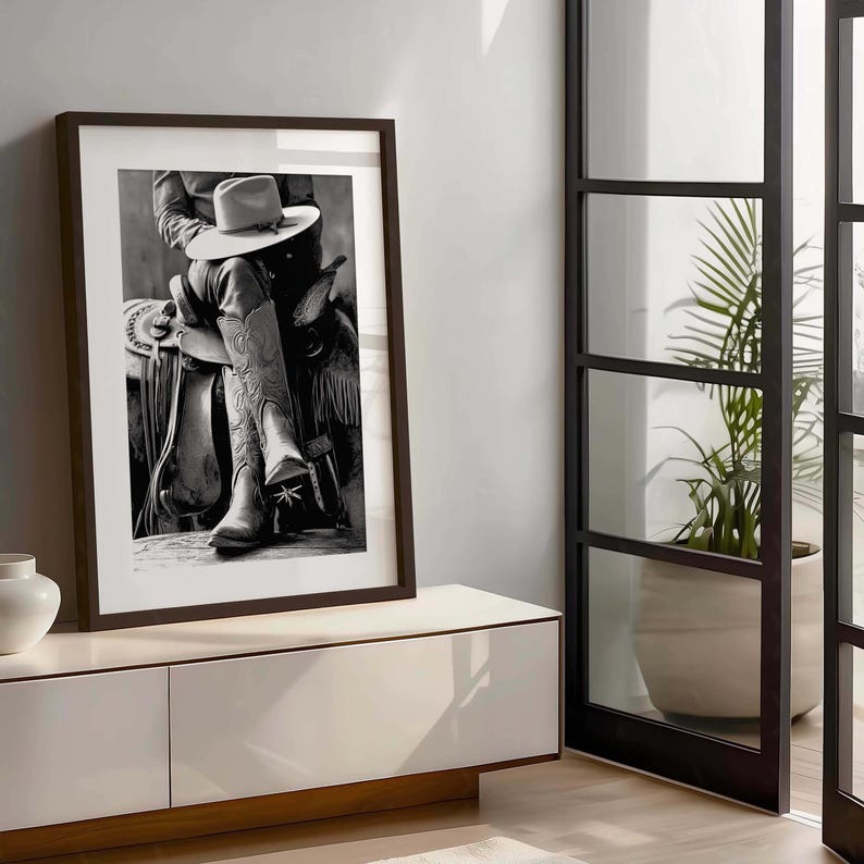 May include: Black and white framed print of a cowboy on a saddle, wearing a hat and cowboy boots. The boots have decorative stitching. The print is displayed on a white cabinet with a brown frame.
