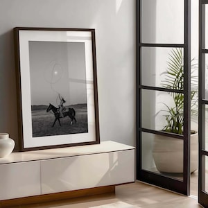 May include: A framed black and white wall art print featuring a person riding a horse in a field while swinging a lasso overhead. The artwork is enclosed in a dark brown wooden frame with a wide white mat, displayed in a vertical orientation.