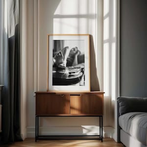 K&ouml;nnte beinhalten: Gerahmtes Schwarz-Wei&szlig;-Foto mit F&uuml;&szlig;en in der N&auml;he eines Plattenspielers. Das Bild wird auf einem Holzschrank mit zwei T&uuml;ren pr&auml;sentiert. Der Schrank steht vor einer wei&szlig;en Wand mit einem Fenster und einem grauen Vorhang.