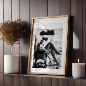 Pode incluir: Impress&atilde;o emoldurada em preto e branco de uma mulher relaxando em uma praia, com um gramofone vintage e uma garrafa em uma pequena mesa de vime. Uma vela acesa e uma planta em vaso est&atilde;o na prateleira abaixo.