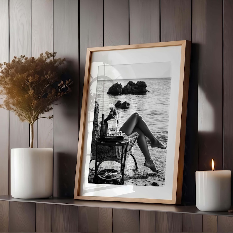 Pode incluir: Impress&atilde;o emoldurada em preto e branco de uma mulher relaxando em uma praia, com um gramofone vintage e uma garrafa em uma pequena mesa de vime. Uma vela acesa e uma planta em vaso est&atilde;o na prateleira abaixo.