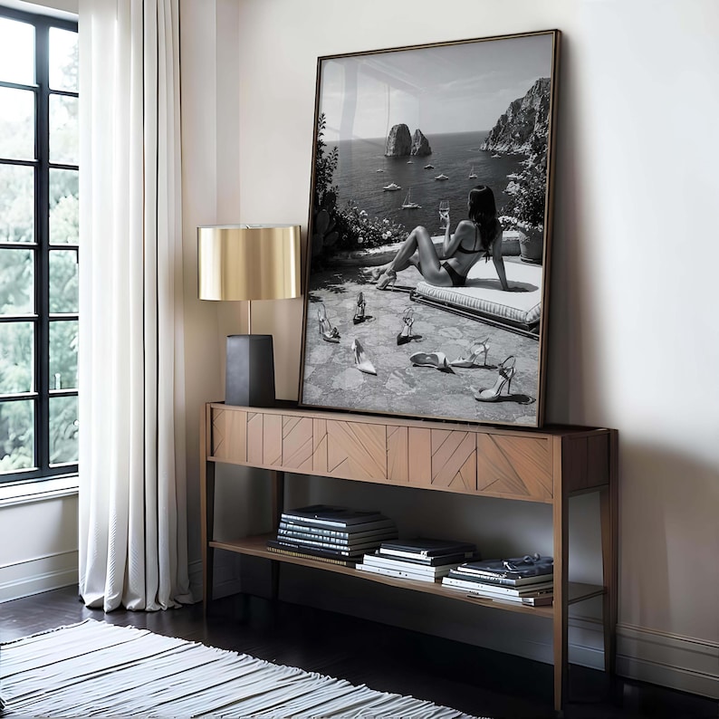 May include: A large rectangular framed wall art piece featuring a black and white photograph of a person in a bikini sitting on a terrace overlooking the sea with several pairs of high-heeled shoes scattered on the stone floor in the foreground.